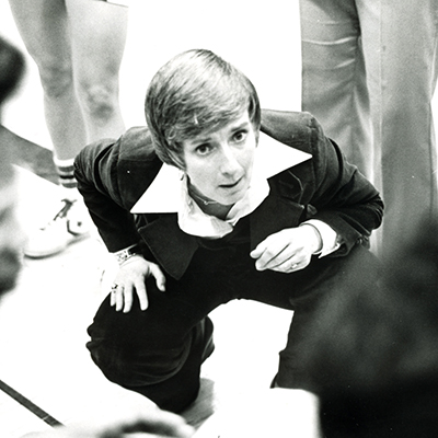 Former women’s basketball and softball head coach Sue (Kruszewski) Hardy ’64 crouches to players in a huddle while coaching a women's basketball game.