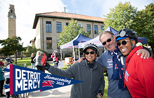 An outdoor photo of three people smiling for the camera, with buildings, trees, bikes and a clock tower in the background.