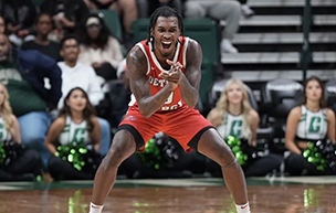 Antoine Davis claps his hands and yells while wearing a red Detroit Mercy uniform. Cheerleaders and other people are pictured behind him inside of an arena.