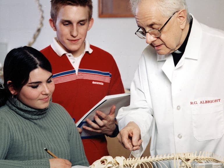 Fr. Albright, wearing a lab coat that bares his name, instructs a pair of students inside of a classroom.