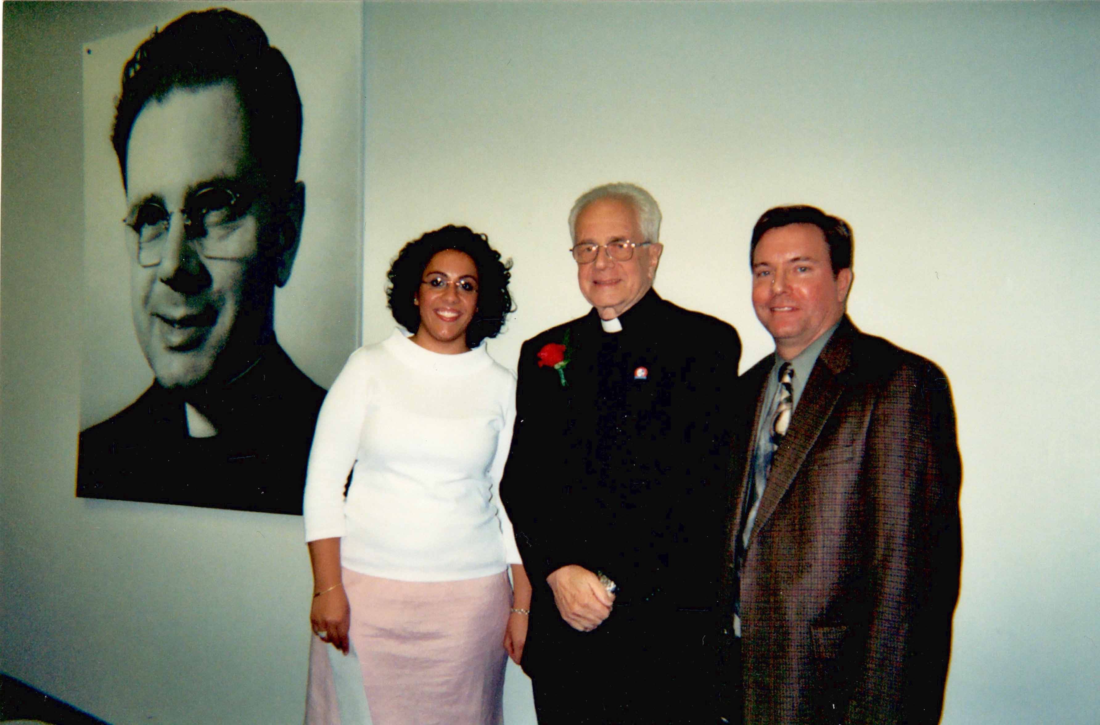 Three people stand in a classroom next to a large photo of Father Albright.