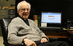Fr. Albright sits in a chair at his desk with a computer in the background.