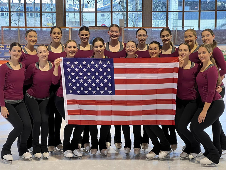 Sixteen skaters stand, holding an American flag on an ice rink.