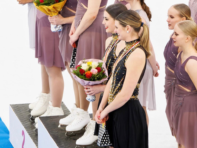 Two skaters stand on a podium, one holding flowers, with other skaters in the background.
