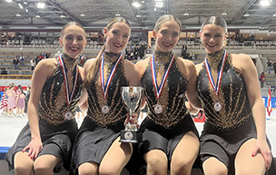 Four skaters wearing medals sit on the boards next to an ice rink with other people behind them.