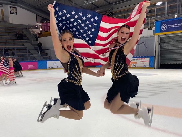 Two skaters leap in the air on an ice rink while holding an American flag.