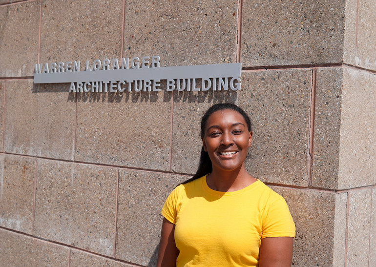 Angel Mangham stands outside next to a brick wall with words on it reading, Warren Loranger Architecture Building.