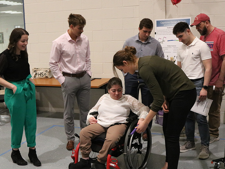 Six people stand and help a recipient, Hope, sitting in her wheelchair, with a Faces on Design design.