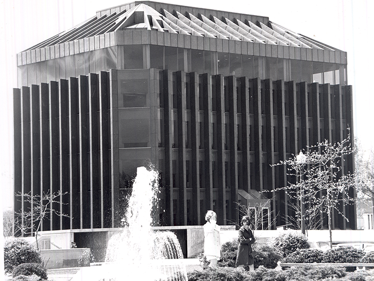 A black and white photo of the Fisher Building, with two people in the foreground walking outdoors near a water fountain.