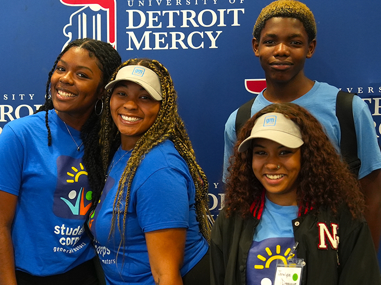 Four high school students stand in front of a blue University of Detroit Mercy backdrop.