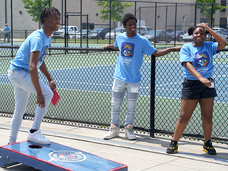 Three high school students wearing Student Corps shirts play corn hole outside near tennis courts.