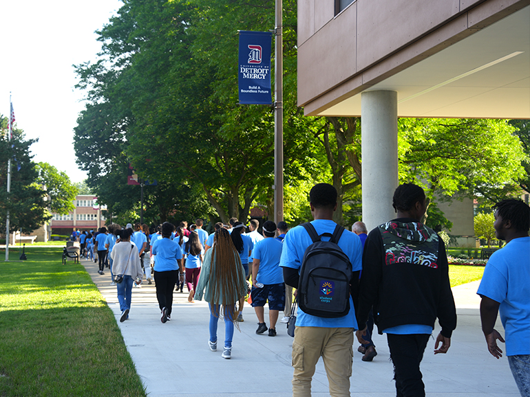 More than three dozen students walk on the sidewalk amongst trees outdoors on the McNichols Campus of Detroit Mercy.