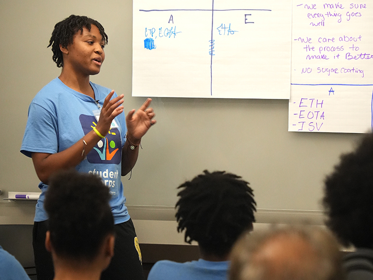 A student speaks near a poster board with words on it inside of a classroom at Detroit Mercy, with others sitting and listening.