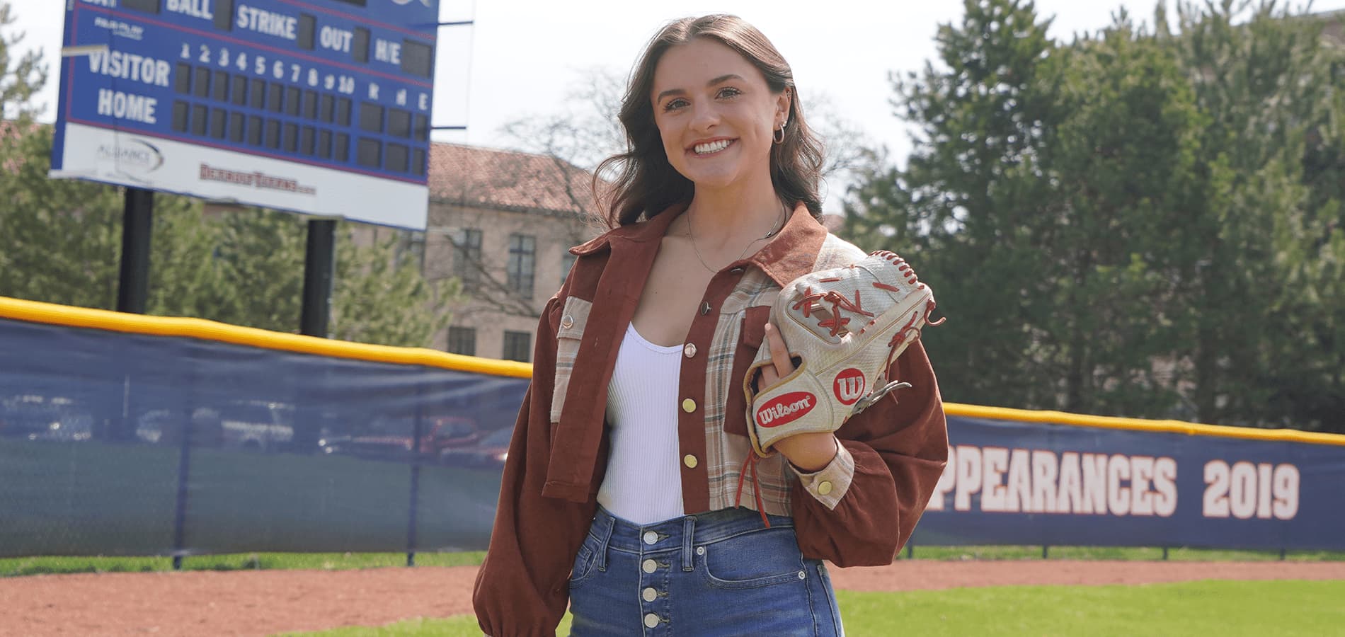 Kara Wolfbauer stands on the Buysse Ballpark field in front of a scoreboard, trees and McNichols Campus buildings on a sunny day.