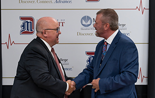 Two men smile and shake hands and look at each other, with logos for University of Detroit Mercy and Macomb Community College on a board behind them.