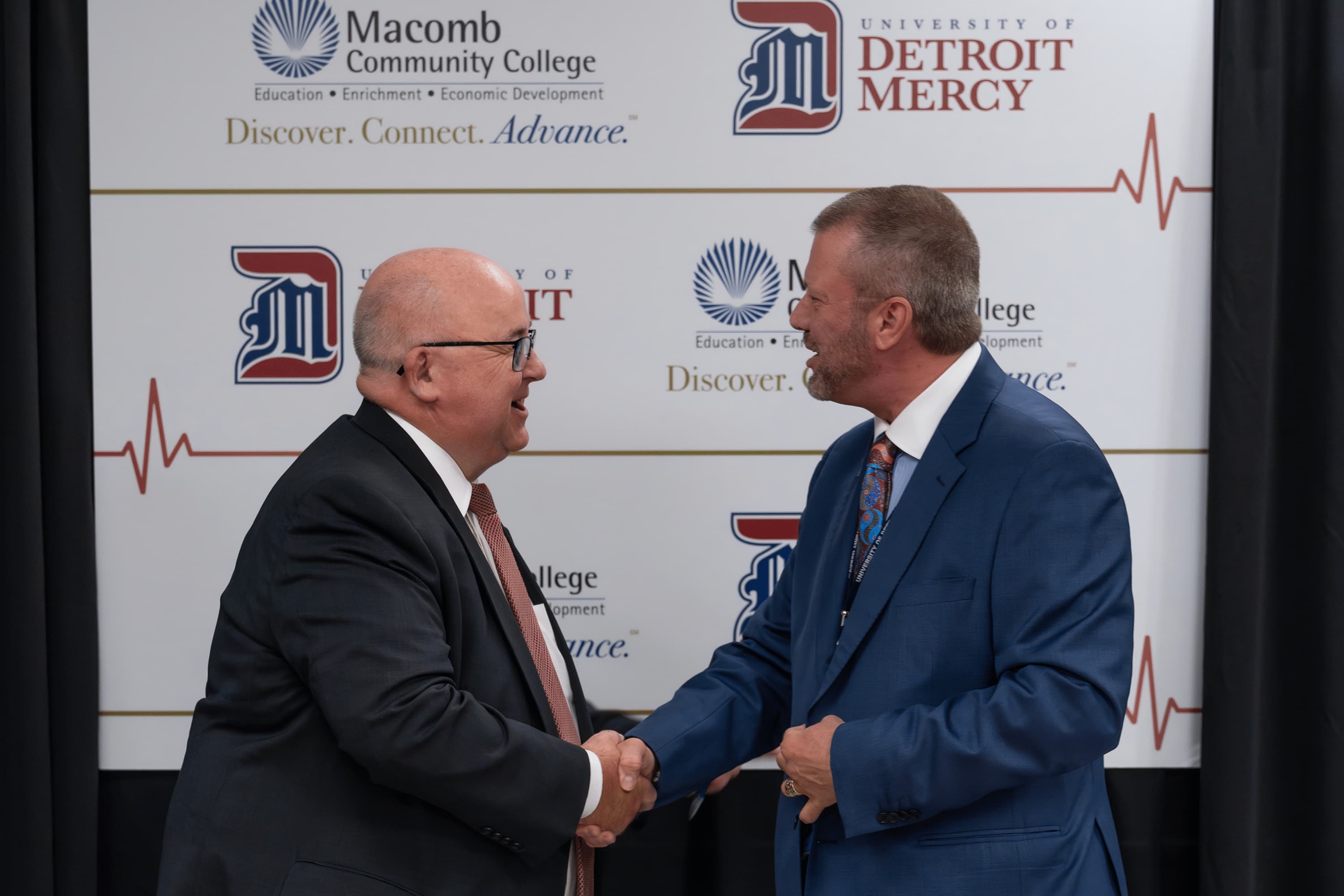 Two men smile and shake hands and look at each other, with logos for University of Detroit Mercy and Macomb Community College on a board behind them.