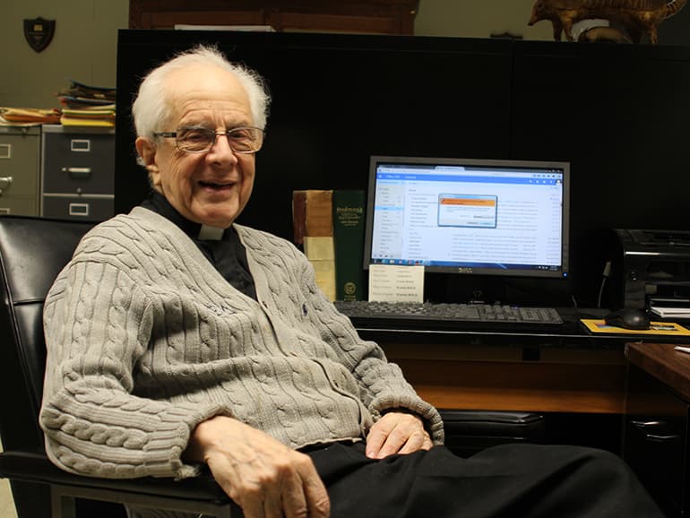Fr. Albright sits at his desk inside of his office, with a computer in the background.