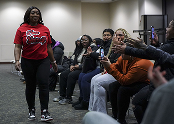 A women wearing a red Pace Yourself t-shirt walks while other sit and watch, with one person holding a phone.