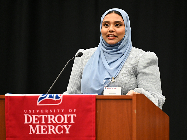 A woman wearing a nametag stands and speaks indoors from a podium, that has a red University of Detroit Mercy banner on it.