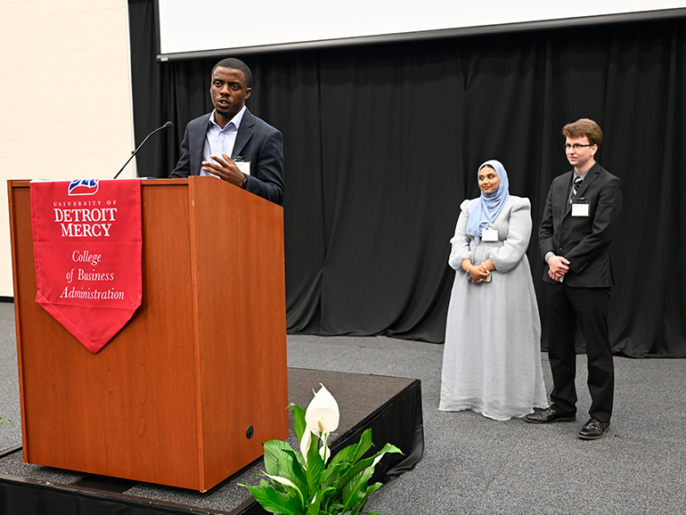 Three students stand inside of the Student Fitness Center, one speaking from a podium that has a red University of Detroit Mercy, College of Business Administration banner hanging from it.