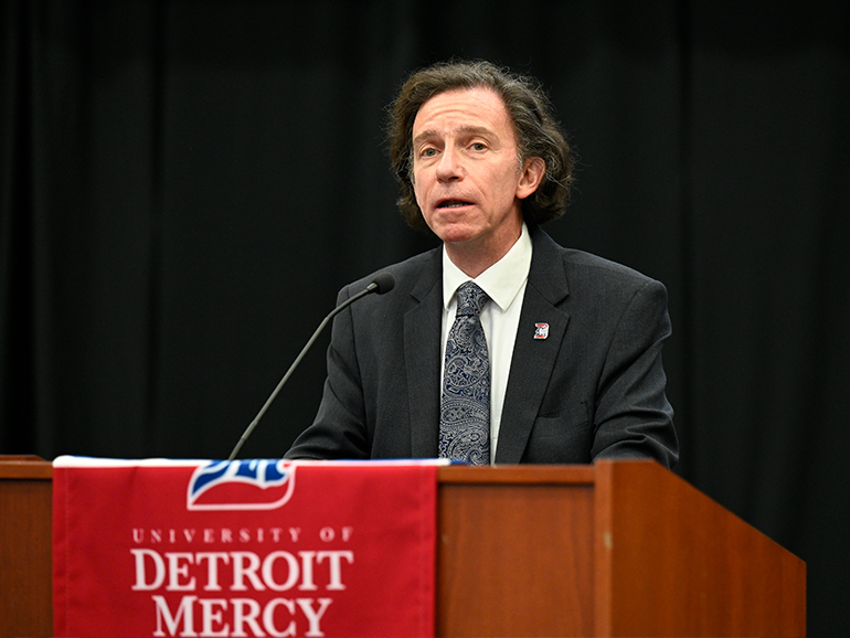 A man wearing a suit stands and speaks indoors from a podium, that has a red University of Detroit Mercy banner on it.