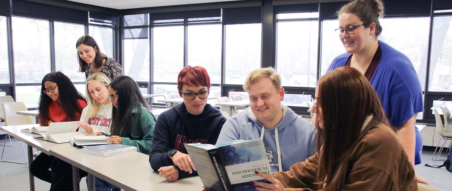 Two groups of three students look at their psychology books while instructors/faculty stand behind them and offer assistance.