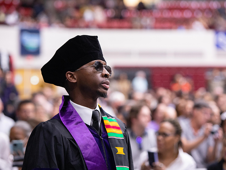 A graduate wearing sunglasses looks on as he waits to walk on stage during commencement.