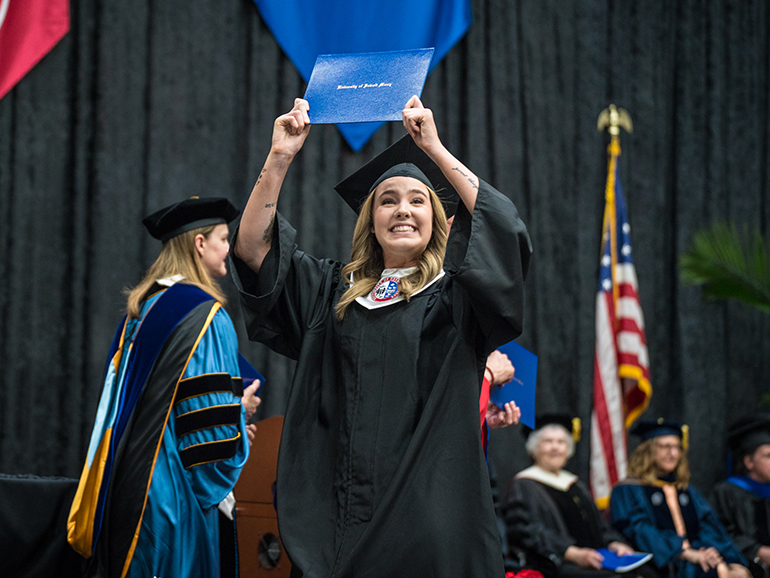 A graduate holds her diploma over her head in celebration as she walks across the stage at commencement.