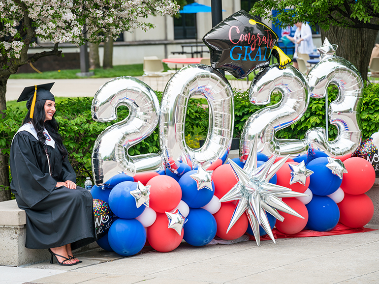 A graduate sits next to a balloon display which features a 2023 and congrats grad balloons and red, white and blue balloons.