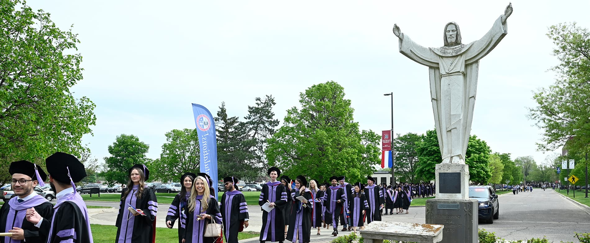 Graduates walk outdoors on the McNichols Campus near the Jesus statue.