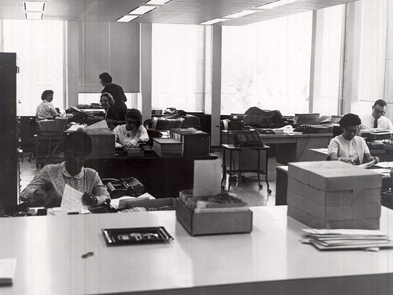 Seven people work at desks in a black and white photo of an office of the Fisher Building.