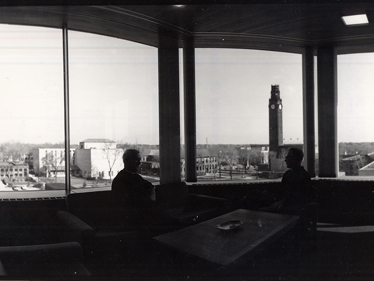 Two people sit inside of an office of the Fisher Building, with University buildings and the clock tower outside of the window.