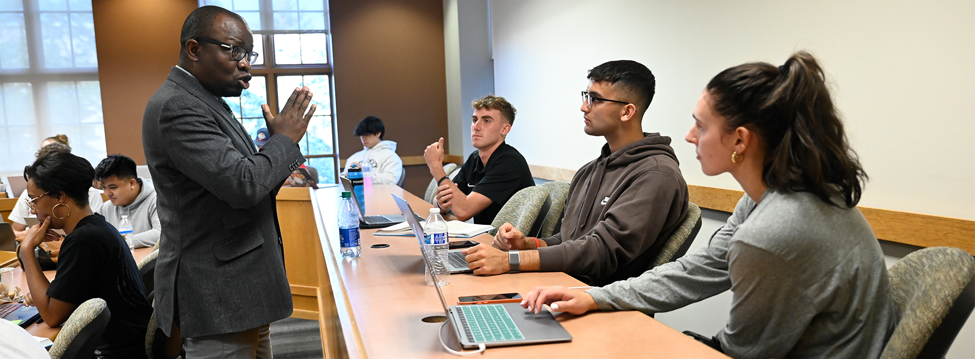 A professor stands and talks to three students sitting in a business classroom. Other students sit in chairs nearby.