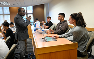 A professor stands and talks to three students sitting in a business classroom. Other students sit in chairs nearby.