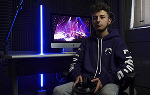 Hisham Almadani holds a gaming controller while sitting in a dark room, with the only light coming from a computer.