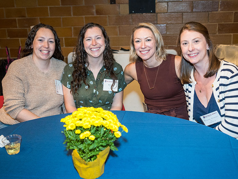 Four women sit around a table and pose for a photo during a Homecoming event. A yellow planted flower is on the table in front of the women.