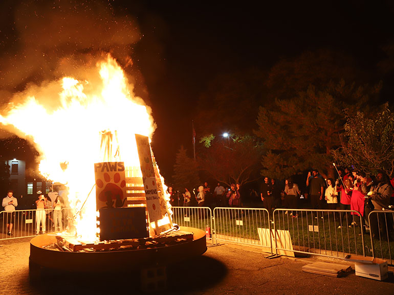 People gather on the grass around a large bonfire in a parking lot. The bonfire features several decorated signs. 