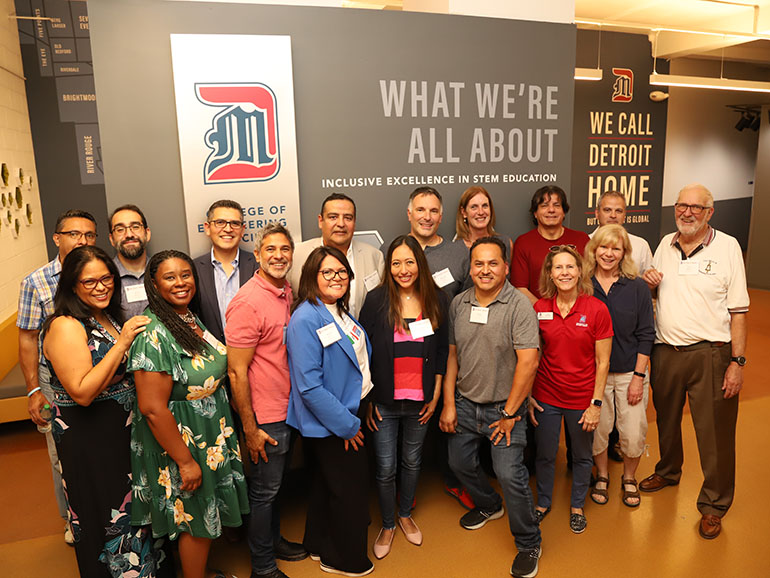 A group of alumni pose for a photo in the College of Engineering & Science's hallway, in front of a decorated wall with several UDM logos and the following text: Detroit Mercy College of Engineering & Science. What we're all about. Inclusive excellence in STEM education. We call Detroit Home. 