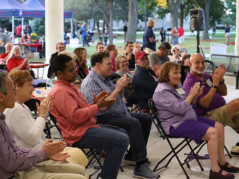 People sitting under the Student Union overhang clap during a Homecoming event.