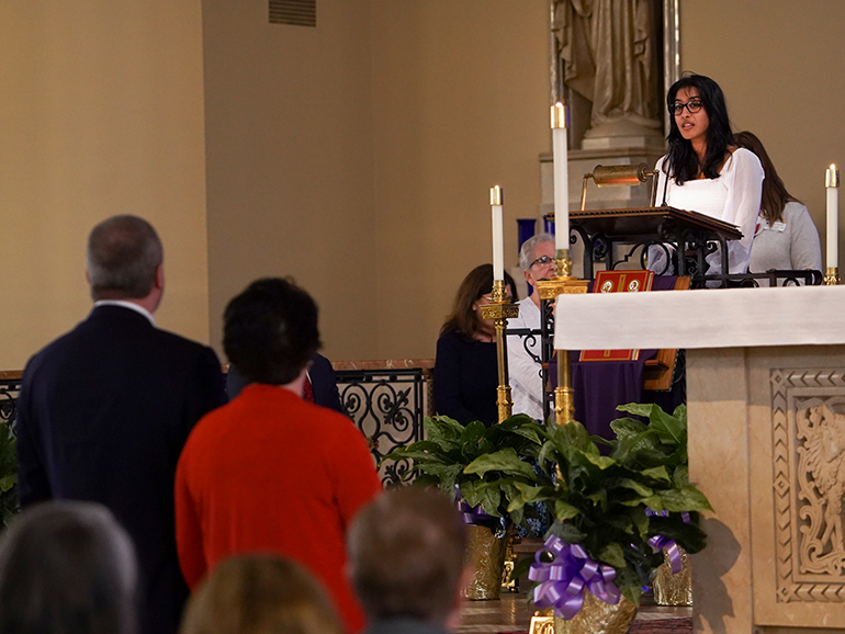 A student speaks from a podium inside of a church, with others pictured in the foreground and background.