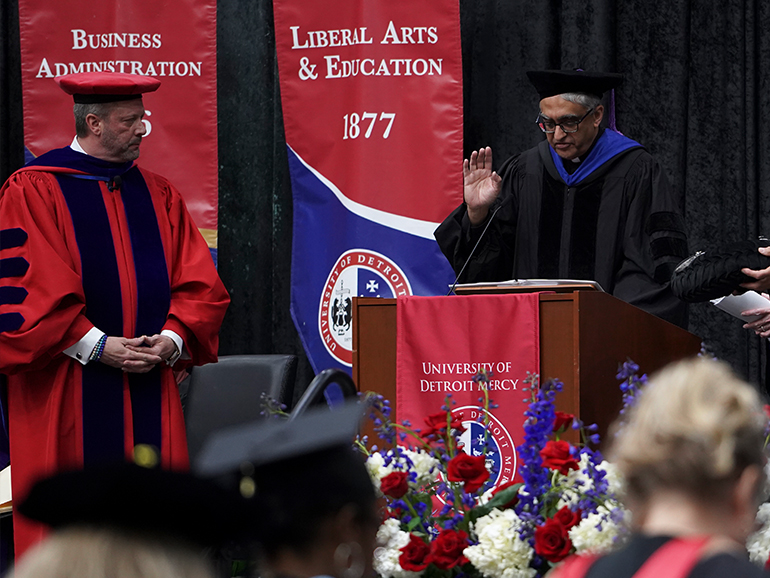 Two people wearing robes stand on a stage with banners on the stage reading, Business Administration, Liberal Arts & Education 1877 and University of Detroit Mercy.