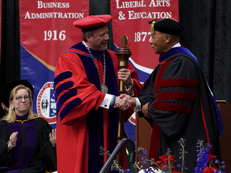 Two people wearing robes shake hands, one holding a mace and an addition person sitting in the background. Banners read Business Administration 1916 and Liberal Arts & Education 1877.