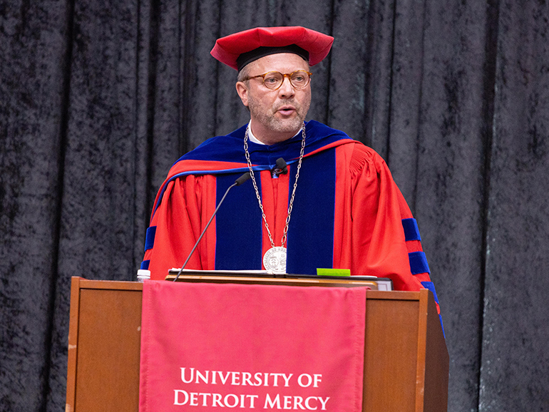 President Taylor speaks from a podium wearing his red robes. A University of Detroit Mercy banner hangs from the podium in front of him.