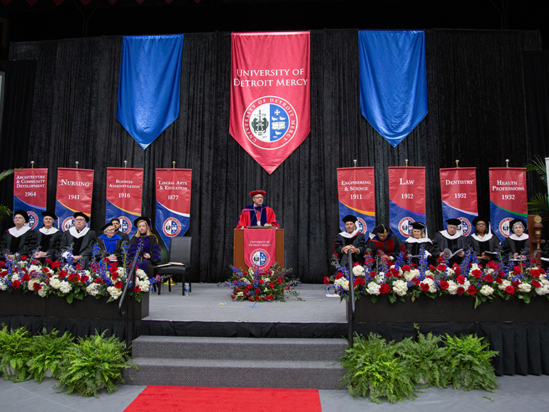 A photo of President Taylor at a podium with people sitting on either side of him and banners of each college behind them. A large University of Detroit Mercy banner hangs behind him.