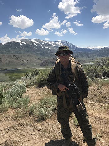 Joe Maki, dressed in his military uniform, is holding a military weapon at his side with a mountain range in the background.