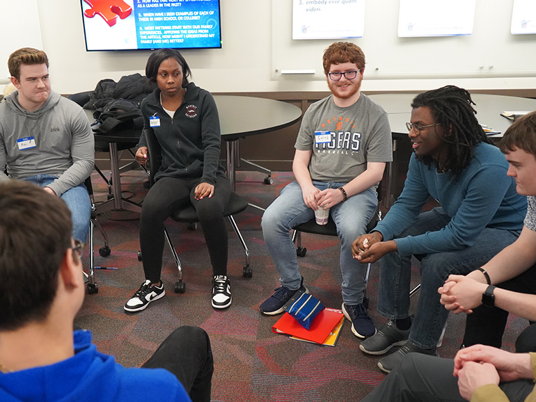 Six students are pictured sitting inside of a classroom during a discussion. One student is wearing a Detroit Tigers t-shirt and text is seen in the background on poster boards.