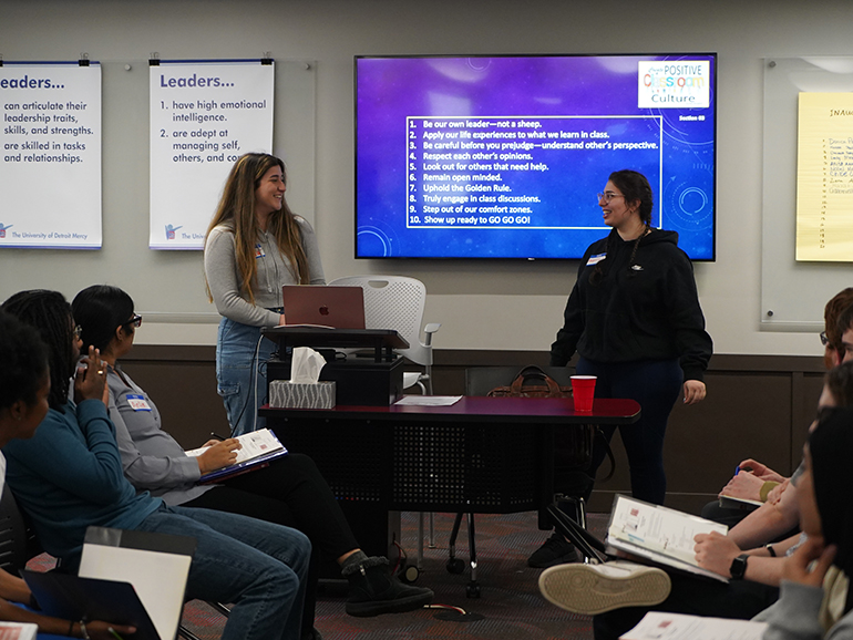 Two students speak at the front of the classroom, with classmates sitting and listening. Leaders posters and a television screen with words on it are in the background.