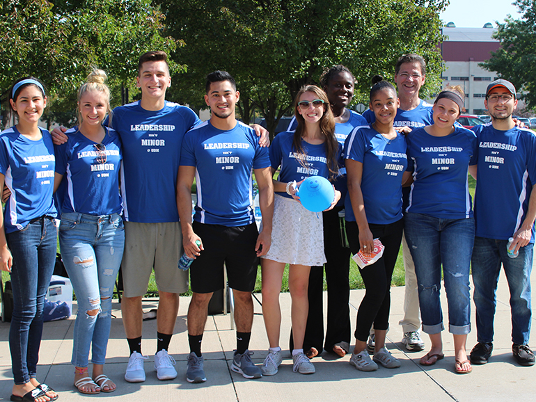 Ten people stand outdoors on the McNichols Campus, all wearing blue Leadership isn't minor at UDM t-shirts.