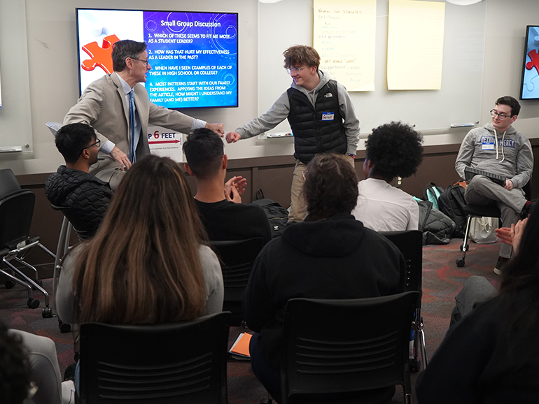 A teacher and student 'fist-bump' inside of a classroom, with several other students sitting and listening. Television screens and poster boards are displayed on the walls.