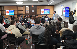 Students sit inside of a classroom and listen to a teacher who sits on a higher chair, with three television screens around them.
